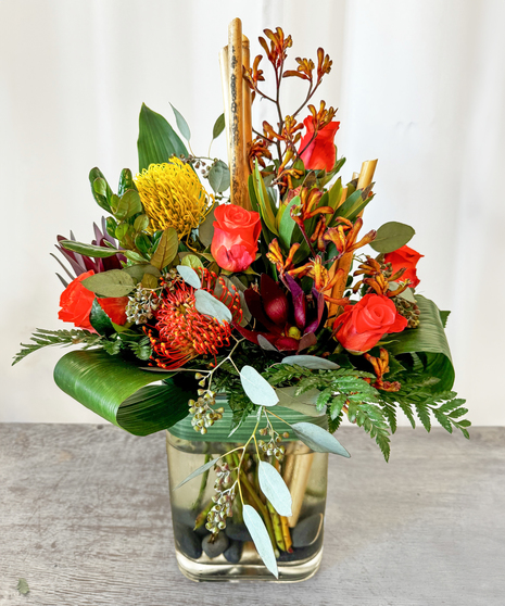 A bright floral arrangement with sunflowers, orange lilies, and coral roses in a turquoise glass vase.